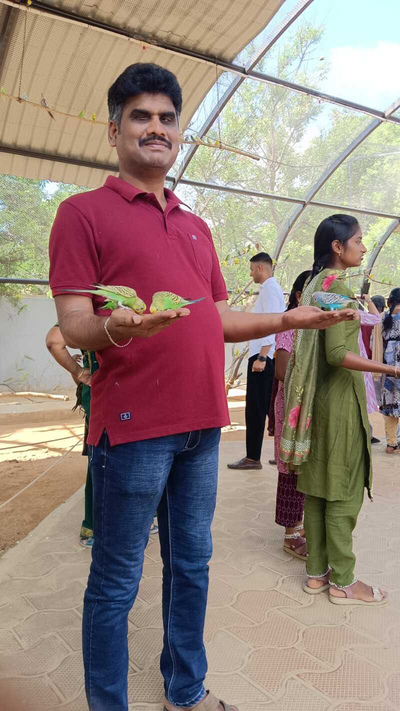 A participant from Thaaikarangal Charitable Trust stands inside the aviary at Rajali Birds Park, Thanjavur, during the field trip, gently holding small parakeets on both hands. Surrounded by fellow students and birds, the vision-disabled participant experiences the joy of connecting with nature through touch, feeling the birds' delicate movements and presence firsthand. A participant from Thaaikarangal Charitable Trust stands inside the aviary at Rajali Birds Park, Thanjavur, during the field trip, gently holding small parakeets on both hands. Surrounded by fellow students and birds, the vision-disabled participant experiences the joy of connecting with nature through touch, feeling the birds' delicate movements and presence firsthand.